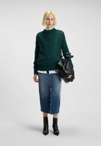 Maglione verde lavorato a maglia, camicia bianca con colletto, gonna a matita in denim, stivaletti neri con punta affusolata, che tiene una borsa nera.