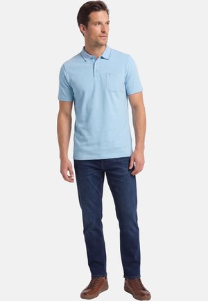 Mann trägt hellblaues Poloshirt, dunkelblaue Jeans und braune Sneakers, steht und schaut seitlich vor weißem Hintergrund.