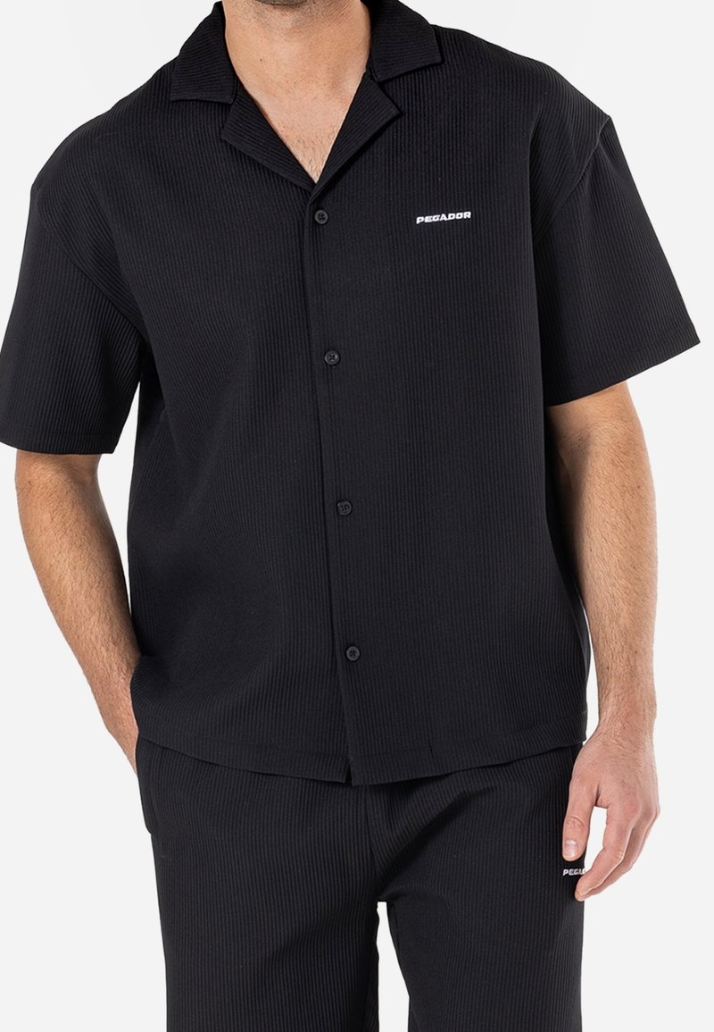 Zwarte korte mouwen shirt van ribmateriaal met een kraag, knoopsluiting aan de voorkant en een klein wit 'PEGADOR' logo aan de linkerkant.