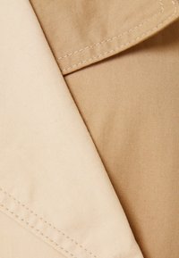 Beżowy materiał o gładkiej fakturze, złożony z widocznymi szwami. Jedna sekcja jest jaśniejsza, podczas gdy druga jest nieco ciemniejszym odcieniem beżu.