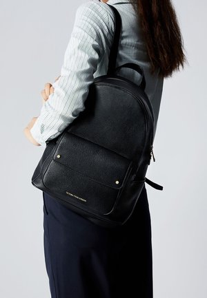 Personne portant un sac à dos en cuir noir avec une poche avant, logo "Victoria Hyde London", chemise rayée et jupe bleu marine sur un fond uni.