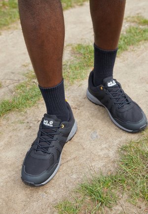 Personne portant des baskets noires Jack Wolfskin avec des chaussettes noires, debout sur un chemin en terre bordé d'herbe.