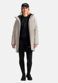 Cappotto isolato beige chiaro con foderatura nera, indossato sopra un maglione nero e leggings. Un cappellino nero con logo è indossato sulla testa.