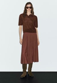 Maglione corto a maniche corte in misto lana marrone con scollo rotondo, abbinato a una gonna plissettata fluida marrone e stivali al ginocchio verde oliva.
