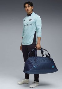 Tmavo modrá cestovná taška s kontrastnými svetlomodrými akcentmi, odolná tkanina, dvojitá rukoväť a zipsová kapsa. Logo zreteľne zobrazené.