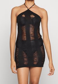 Donna che indossa un mini abito nero senza maniche con pannelli di maglia strappati verticali che rivelano la pelle e la biancheria intima sottostante.