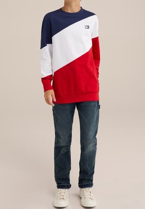 Garçon portant un t-shirt à manches longues color-block bleu marine, blanc et rouge, un jean foncé et des baskets blanches sur un fond neutre.