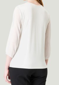 Haut blanc avec manches transparentes trois-quarts, encolure ronde et finition douce et texturée. Porté avec un pantalon noir.