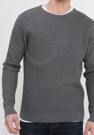 Grau strukturierter Strickpullover, getragen über einem weißen Unterhemd, kombiniert mit dunkelblauen Jeans, gezeigt an einer stehenden Person vom Hals bis zur Hüfte.