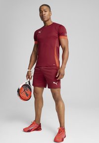 Raudona sportinė marškinė ir šortai su oranžiniais akcentais, derinami su raudonais sportiniais batais ir laikant juodą bei raudoną raketę ekstremaliam tenisui.