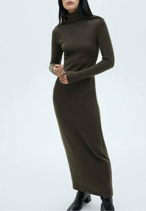 Femme portant une longue robe ajustée col roulé olive foncé avec manches longues et des bottines noires, debout devant un fond blanc.