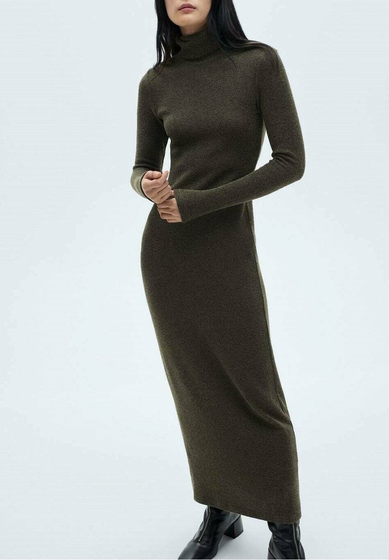 Femme portant une longue robe ajustée col roulé olive foncé avec manches longues et des bottines noires, debout devant un fond blanc.