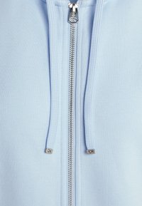 Ljusblå zip-up hoodie med en slät, texturerad tyg, silverdragkedja och märkta dragkedjeöglor med "CK"-detaljer.