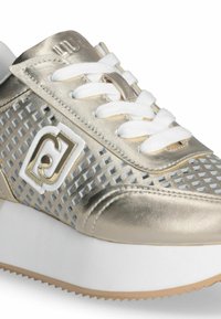 Metallic auksinės spalvos sportbačiai su perforuotomis dalimis, baltais raišteliais ir baltu logotipu šone, turintys storą baltą padu ir tekstūrinius akcentus.