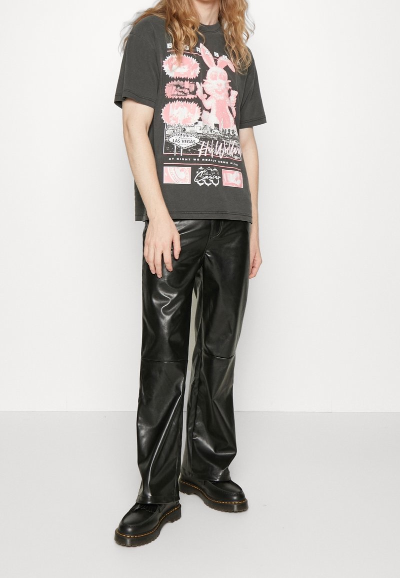 Personne portant un t-shirt noir avec un imprimé lapin rose, un pantalon en cuir synthétique noir et des chaussures plateformes noires, debout sur fond blanc.
