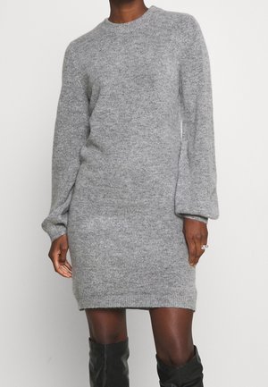 Personne portant une robe-pull tricotée gris clair à manches longues, longueur genoux, avec des bottes noires jusqu’aux genoux, debout devant un fond uni.