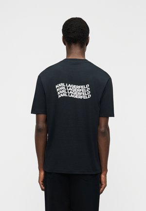 Man seen from behind wearing a black T-shirt with “KARL LAGERFELD” repeated four times in white text on the upper back.