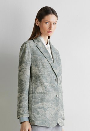 Frau mit glattem braunem Haar trägt ein graues, mit Blumenmuster versehenes Blazer über einem weißen Hemd und steht vor einer schlichten hellen Wand.