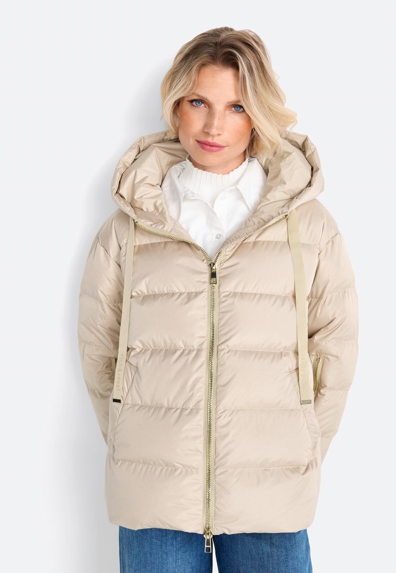 Beige pufferjack met een capuchon, vooraan een rits en een verticale gestepte design. Bevat verstelbare banden en twee zijzakken. Heeft een soepele textuur.