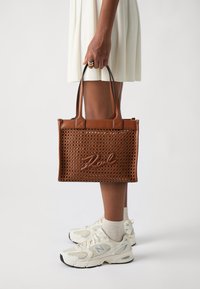 Borsa tote intrecciata marrone con scritte in rilievo, doppio manico e base a tinta unita contrastante. Abbinata a sneakers bianche e un vestito leggero.