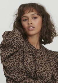 Haut à motif léopard marron avec des manches bouffantes, présentant une texture lisse et un col rond. Le motif comprend diverses nuances de marron et de noir.