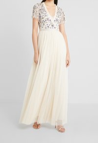 Maxikleid in Creme mit V-Ausschnitt, kurzen bestickten Ärmeln und floralen Verzierungen. Fließender Tüllrock mit geschichteter Textur.