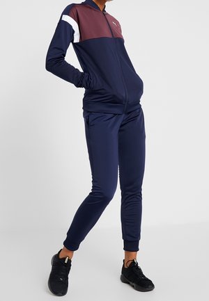 Persona que lleva un chándal azul marino con panel superior burdeos, raya blanca, zapatillas negras, de pie contra un fondo claro liso, manos en los bolsillos.