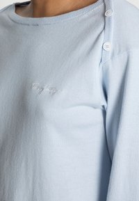 Hellblaues Strickoberteil mit zwei weißen Knöpfen an der Schulter und kleinem bestickten, geschwungenen Text auf der Brust.