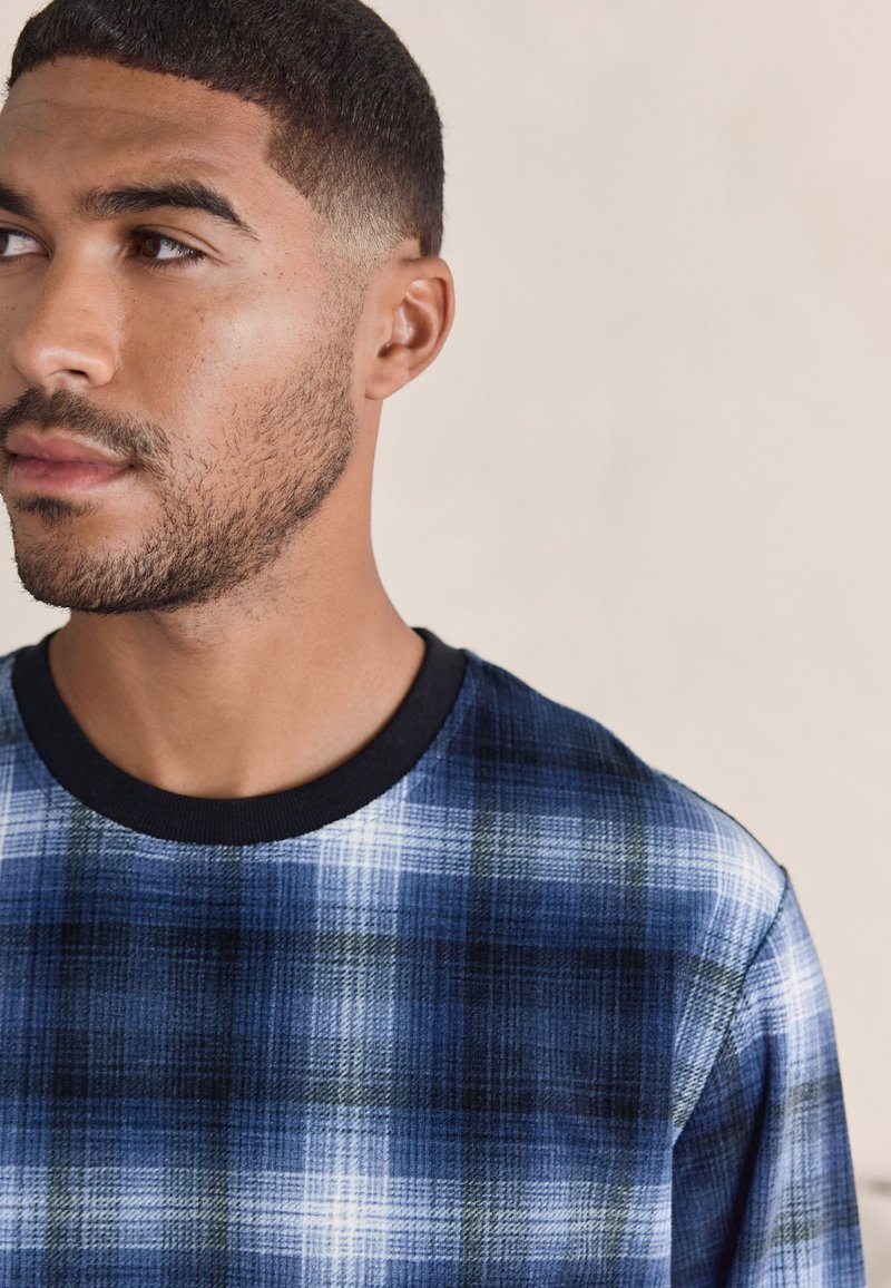 Bărbat tânăr cu păr scurt și barbă, purtând o cămașă în carouri albastre și negre, uitându-se spre stânga pe un fundal bej.