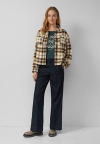 Geruite jas in crème, bruin en zwart; groene graphic t-shirt; donkerblauwe wijde broek; taupe schoenen met gestructureerde zolen.