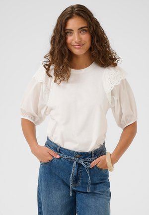 Top bianco a maniche corte con dettagli in pizzo ricamato smerlato sulle spalle. Abbinato a pantaloni di jeans blu a vita alta, con cintura annodata.