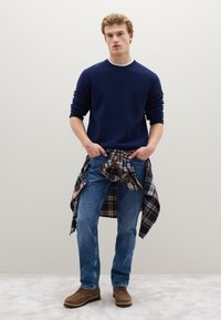 Marineblauer Strickpullover, blaue Jeans, braune Stiefel und ein braun kariertes Hemd, das in der Taille gebunden ist. Einfaches Design, lässige Passform, strukturierter Stoff.