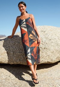 Abito lungo con spalline sottili, caratterizzato da un motivo geometrico colorato e foglie di palma in arancione, nero e crema su uno sfondo scuro.