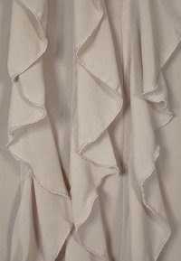 Beżowy materiał z falbaniastymi krawędziami i srebrnym przeszyciem. Tekstura wydaje się miękka, a warstwowe fałdy tworzą płynny wzór.