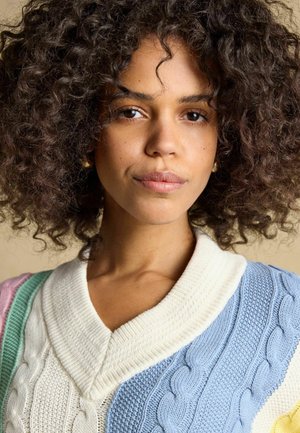 Young woman with curly hair wearing a pastel-colored, multi-textured cable knit sweater with a white collar, looking confidently ahead.