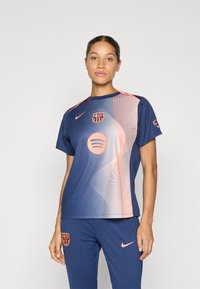 Woman standing in blue and pink FC Barcelona sportswear with Nike logos and Spotify emblem on plain white background.