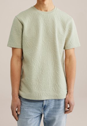 Chemise à manches courtes vert clair texturée avec des motifs abstraits en relief portée par une personne en jean bleu clair.