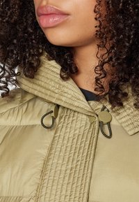 Khaki puffer bunda s jemne prešívanou textúrou, veľkou kapucňou a sťahovacími šnúrkami. Disponuje hladkým materiálom a subtilným brandingom.