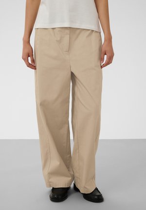 Personne portant un pantalon beige ample, un haut blanc et des chaussures noires se tenant sur un sol gris clair devant un fond uni.