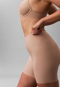 Vrouw die hoog getailleerde naadloze nude shapewear-shorts en bijpassende bh aanpast tegen een neutrale grijze achtergrond.