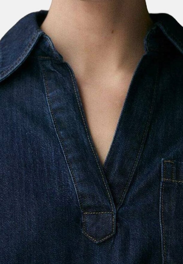 Gros plan d’une personne portant une chemise en denim bleu foncé avec un col en V et des détails de couture visibles.