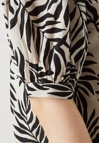 Μαύρη και κρεμ μπλούζα με σχέδιο φύλλων, διαθέτει συγκεντρωμένα μανίκια και λεπτομέρεια στον σφιγκτήρα. Κατασκευασμένη από ελαφρύ ύφασμα με λεία υφή.