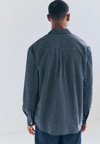 Camisa de hombre de manga larga con un patrón de cuadros en azul marino y blanco. Presenta un collar clásico y puños abotonados. Textura sutil, detalle en el yugo trasero.