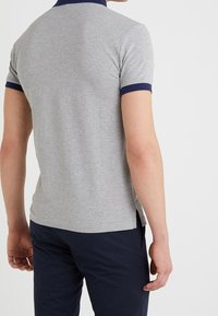 Grå bomullspolo med marinblå krage och ärmkanter, figursydd design, sidoflikar och en slät yta, sedd bakifrån.