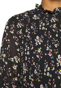 Blouse noire plissée avec col montant à volants, manches transparentes et petit motif floral multicolore en blanc, jaune, rouge et bleu.