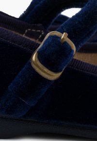 Sándalo de terciopelo azul oscuro con una correa a juego y un hebilla de metal dorado. La superficie es suave con un ligero brillo y suela negra resistente.