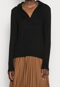 Černý pulovr s dlouhými rukávy, kapucí a výstřihem do V. Měkká, hladká textura; nošený přes plisovanou hnědou sukni, která zvýrazňuje jeho vrstvený vzhled.