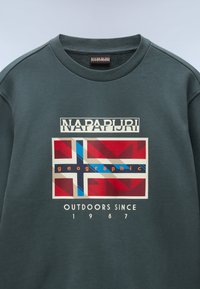 Zelená mikina s žebrovaným kulatým výstřihem. Obsahuje centrální grafický design s motivem vlajky, text "NAPAPIJRI" a "OUTDOORS SINCE 1987."