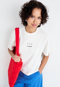 Hvid bomulds t-shirt med sort "CPH STUDIOS" tekst, blå bukser og en rød quiltet tote bag. Blød tekstur og løstsiddende pasform.
