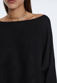 Pull noir à épaules dénudées en maille côtelée, avec une coupe ample et un tissu texturé. Un collier ras du cou fin en métal est superposé autour du décolleté.